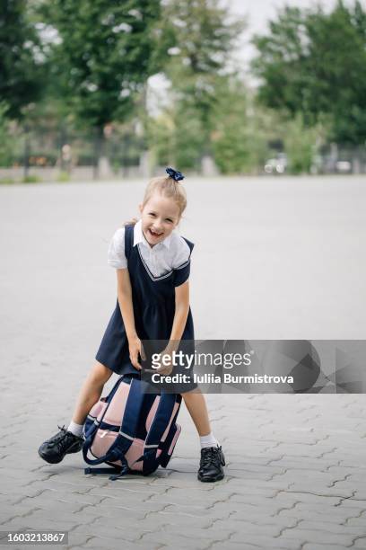 young blonde girl in blue school uniform standing in schoolyard holding satchel and smiling - school kids stock pictures, royalty-free photos & images