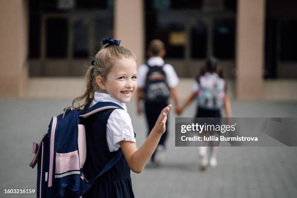 photo of pre-adolescent blonde girl in school uniform with backpack looking back smiling and waving - first day of school fotografías e imágenes de stock