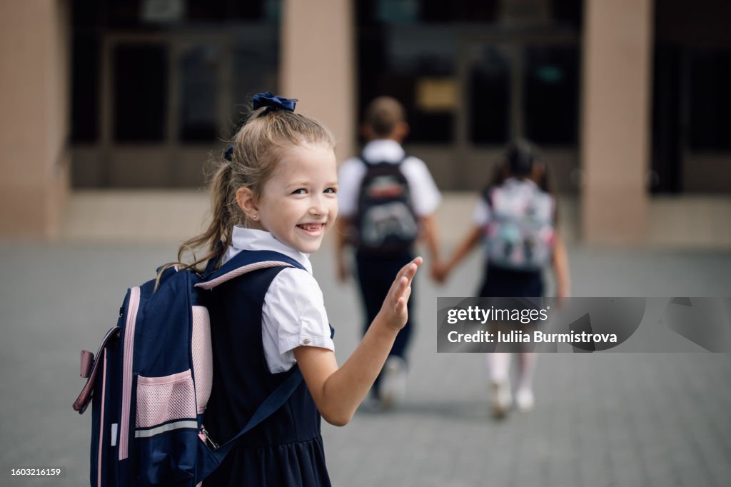 Photo of pre-adolescent blonde girl in school uniform with backpack looking back smiling and waving