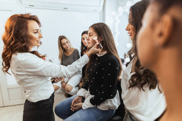 Woman in front of students, applying make-up to another woman who is sitting in a chair.