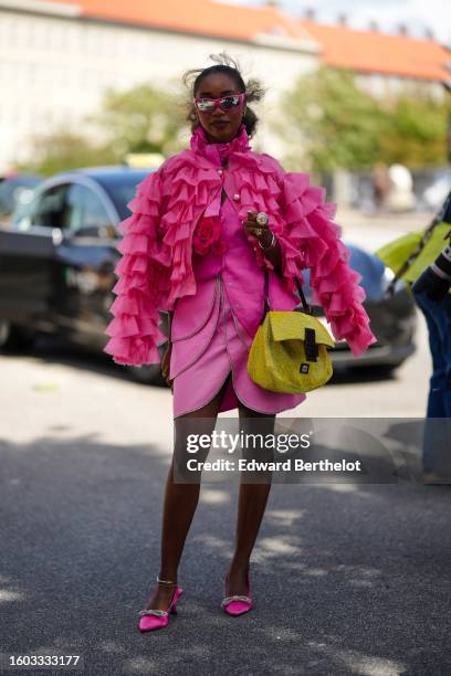 Guest wears neon pink sunglasses, earrings, a neon pink ruffled high neck / long sleeves cropped silk satin blouse, a silver watch, gold rings, a...