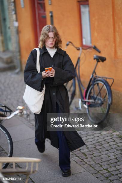 Guest wears a white latte turtleneck pullover, a black buttoned long coat, a white suede large shoulder bag, navy blue denim wide legs pants, black...