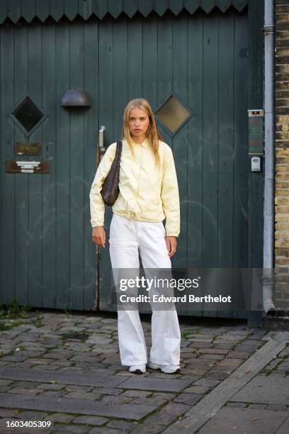 Guest wears a pale yellow zipper jacket, a dark brown braided shiny leather shoulder bag, white denim wide legs pants, white sneakers , outside...