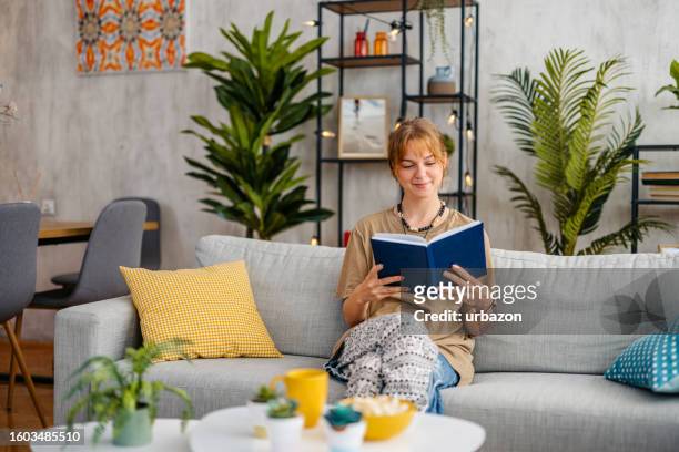 donna che si siede sul divano e legge un libro a casa - sfogliare libro foto e immagini stock