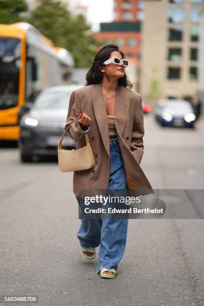 Guest wears white large squared sunglasses, gold chain pendant necklace, a brown beige oversized blazer jacket, a multicolored braided wool striped...