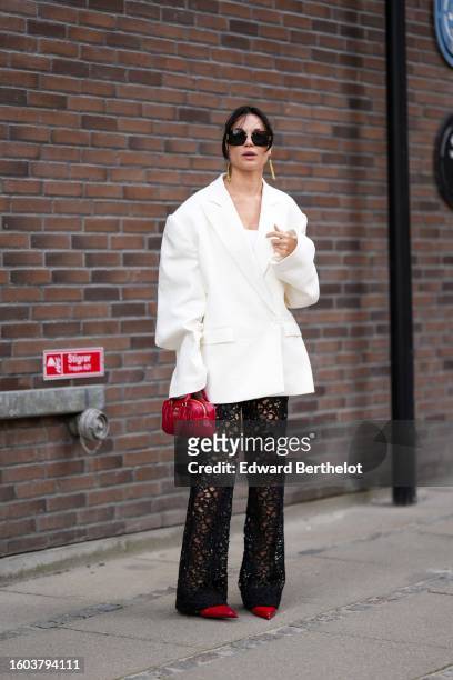 Guest wears black sunglasses, gold pendant earrings, a white oversized blazer jacket, a white shoulder-off top, black lace cut-out pattern large...