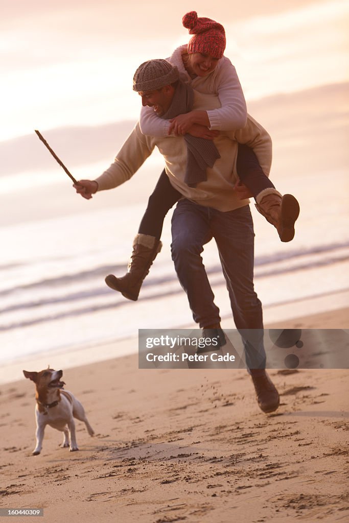 Couple piggyback on beach in winter with dog