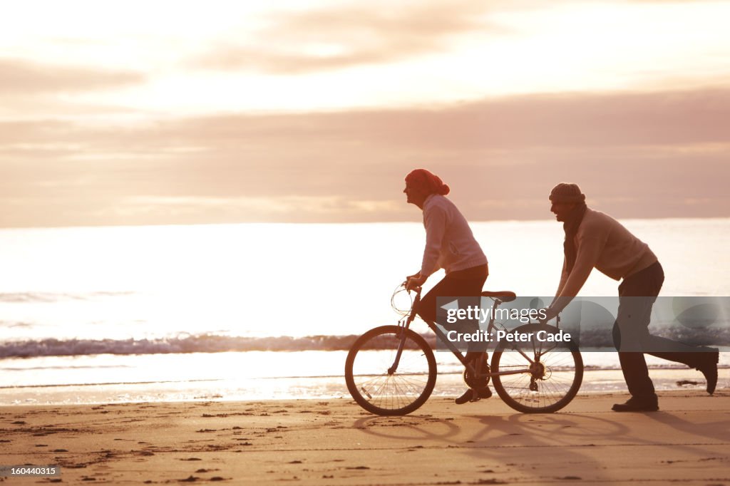 Man pushing woman on bike, on beach a sunset