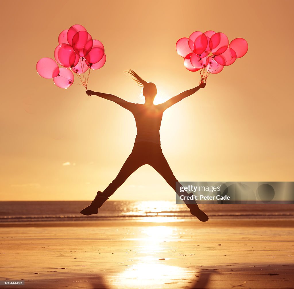 Woman jumping on beach at sunset holding balloons