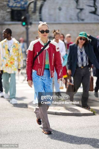 Guest wears a white ribbon hair elastic, black sunglasses, a green and yellow braided wool t-shirt, a red and white shiny leather zipper jacket, a...