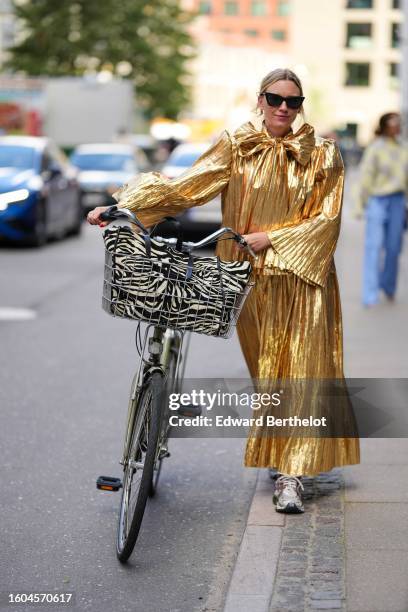 Guest wears black butterfly sunglasses, a gold shiny varnished pleated / accordion flared long sleeves / knot bow tie neck / long dress, a black and...