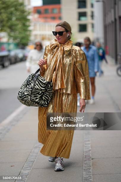 Guest wears black butterfly sunglasses, a gold shiny varnished pleated / accordion flared long sleeves / knot bow tie neck / long dress, a black and...