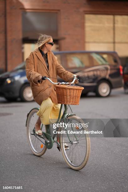 Guest wears black sunglasses, a dark brown shiny leather shirt, a camel oversized blazer jacket, a pale yellow silk skirt, white latte leather block...