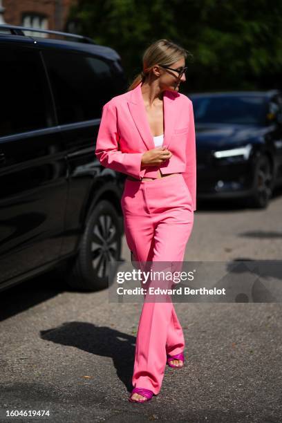 Guest wears black sunglasses, a white t-shirt, a neon pink cropped blazer jacket, matching neon pink flared suit pants, neon pink shiny leather...