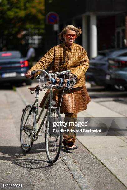 Guest wears red futurist sunglasses, a camel quilted rain coat, a matching dark brown quilted rain long skirt, matching gold quilted rain pants,...