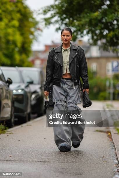 Gili Biegun wears a black shiny leather zipper biker jacket, a pale khaki denim cropped shirt, gray denim laces knees large wide legs pants, a black...