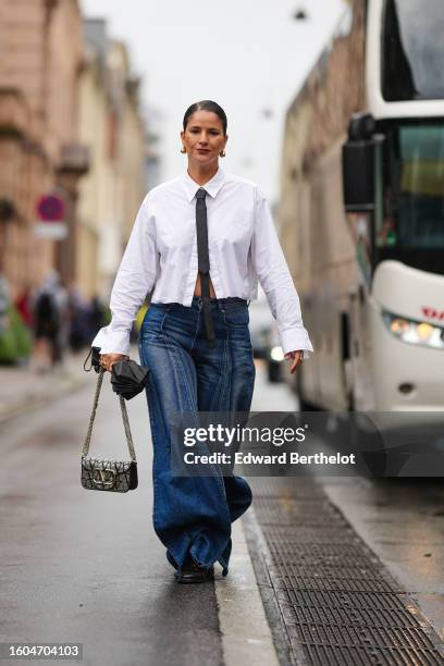 Gili Biegun wears gold pendant earrings, a black tie, a white shirt, navy blue embossed faded pattern puffy large baggy pants, a black umbrella, a...