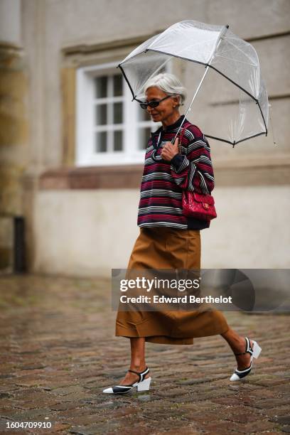 Grece Ghanem wears a white transparent umbrella, black sunglasses, a white t-shirt, a red / black / white striped print pattern zipper neck pullover,...