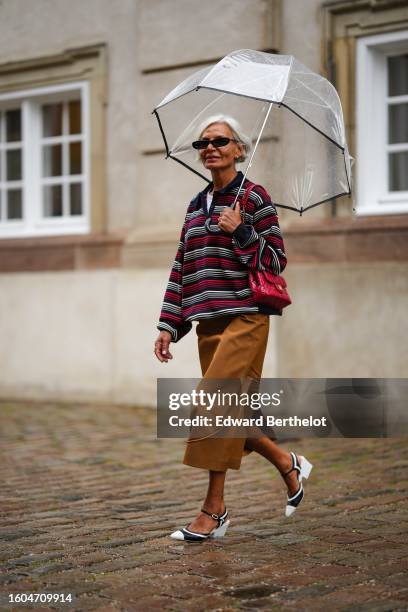 Grece Ghanem wears a white transparent umbrella, black sunglasses, a white t-shirt, a red / black / white striped print pattern zipper neck pullover,...