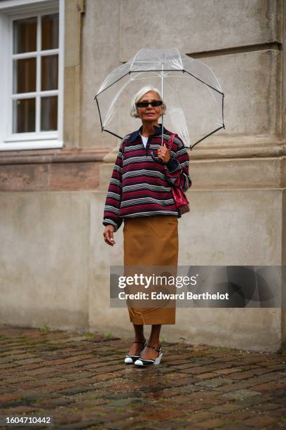 Grece Ghanem wears a white transparent umbrella, black sunglasses, a white t-shirt, a red / black / white striped print pattern zipper neck pullover,...