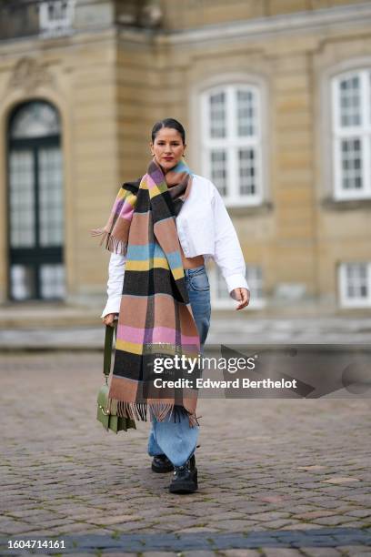 Gili Biegun wears a white cropped open shirt, a brown / black / yellow / blue / pink large striped print pattern fringed wool scarf, a khaki matte...