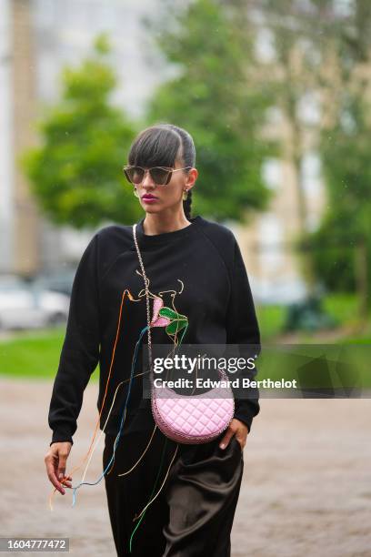 Katya Tolstova wears black sunglasses, green earrings, a black with embroidered multicolored fringed pattern sweater, a pale pink shiny leather...