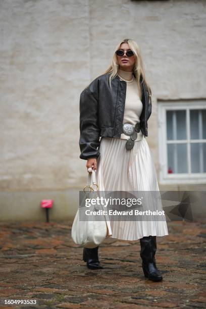 Guest wears black sunglasses, a beige ribbed wool turtleneck pullover, a white pearls necklace, a black shiny leather bomber coat, a dark gray matte...