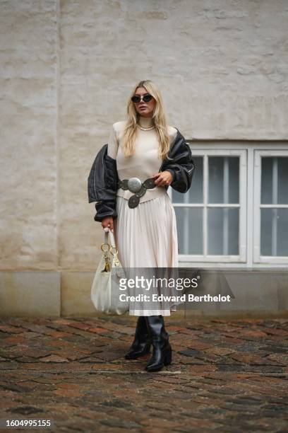 Guest wears black sunglasses, a beige ribbed wool turtleneck pullover, a white pearls necklace, a black shiny leather bomber coat, a dark gray matte...