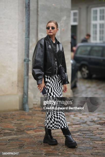 Guest wears black sunglasses, a black pullover, a black shiny leather bomber coat, a black and white asymmetric checkered print pattern long dress,...