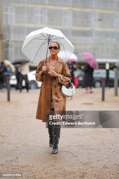 Guest wears a white transparent umbrella, black sunglasses, a camel shiny leather long coat, gold necklaces, a beige braided wool short dress from...