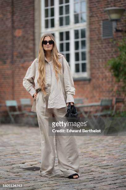 Guest wears black sunglasses, a gold Moove necklace from Messika, a beige denim oversized shirt, matching beige denim wide legs pants, a black...