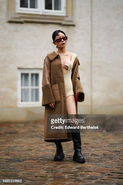 Guest wears black and orange squared sunglasses, a beige heart-neck / cut-out chest / short tube jumpsuit, a brown shiny leather and sheep fur long...