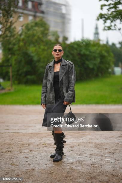 Guest wears black and silver sunglasses, gold earrings, a black small silk scarf, a gold chain necklace, a black shiny leather oversized blazer...