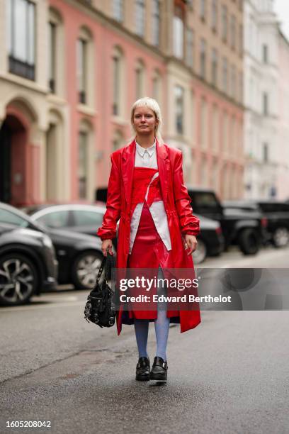 Guest wears silver earrings, a white shirt, a neon red shoulder-off lace print pattern cropped corset top, a red shiny leather trench coat, a red...