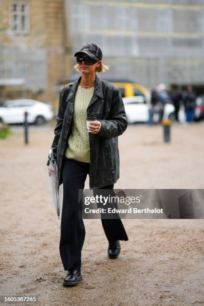 Guest wears a black denim with white embroidered pattern cap, black sunglasses, a gold large chain necklace, a pale green wool pullover, a black...