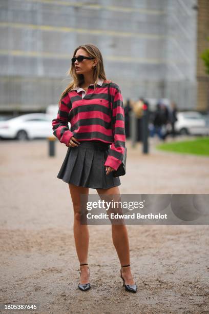 Guest wears black sunglasses, a red and gray striped print pattern polo shirt from Ralph Lauren, a dark gray denim pleated / accordion short skirt, a...