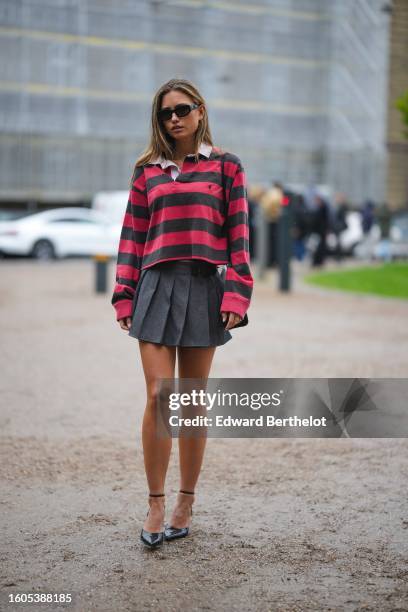 Guest wears black sunglasses, a red and gray striped print pattern polo shirt from Ralph Lauren, a dark gray denim pleated / accordion short skirt, a...