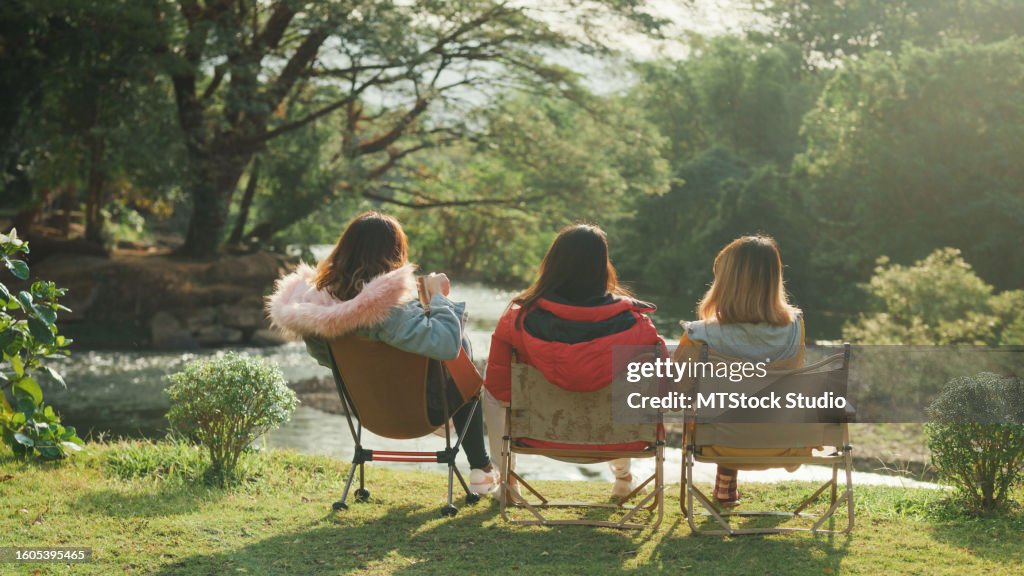 Group of happy Asian friends talking and drinking a cup of hot coffee for breakfast near stream in vacation with camping caravan at morning. Lifestyle travel nature.