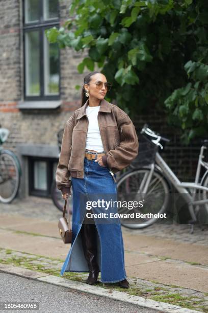 Guest wears brown sunglasses, a white t-shirt, a dark brown faded leather bomber coat, a brown leather belt, a navy blue denim slit / split long...