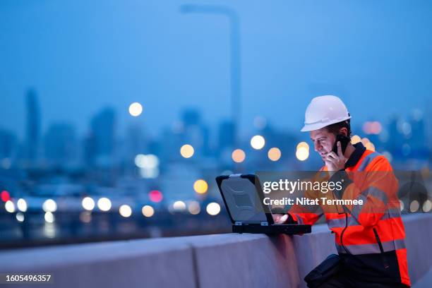 improved communication and collaboration with construction management software for project real-time insights. a project manager in a suspension bridge construction site while using a laptop computer and discussing project tasks with his team via phone. - hybrid cloud stock pictures, royalty-free photos & images