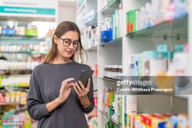 young woman smiling while using smartphone standing against shelf in pharmacy - verschreibungspflichtiges medikament stock-fotos und bilder