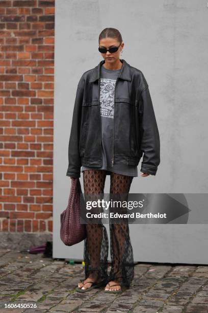 Guest wears black sunglasses, a gray with white print pattern oversized t-shirt, a black shiny leather bomber jacket, black lace print pattern tulle...