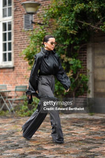 Guest wears gold large futurist sunglasses, gold earrings, a black high neck silk / satin long sleeves blouse, a black shiny leather belt, gray...