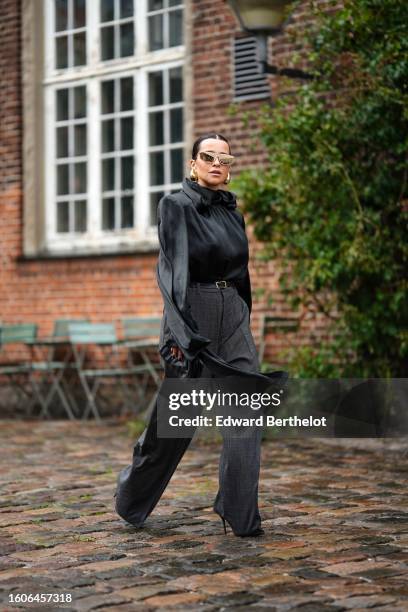 Guest wears gold large futurist sunglasses, gold earrings, a black high neck silk / satin long sleeves blouse, a black shiny leather belt, gray...