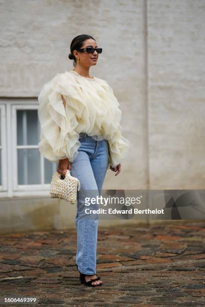 Guest wears black sunglasses, a pale yellow oversized tulle ruffled long sleeves blouse, pale blue denim large pants, a pale yellow and white braided...