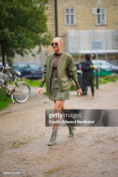 Guest wears black aviator sunglasses, a black lace print pattern long sleeves t-shirt, a khaki quilted jacket, a khaki short skirt, white socks, pale...