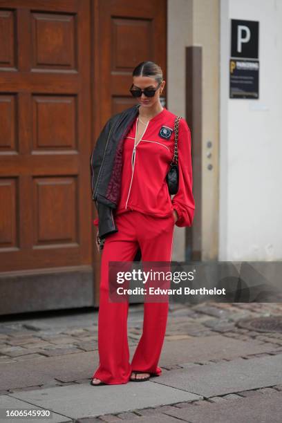 Guest wears black sunglasses, gold earrings, gold necklaces, a black shiny leather jacket, a flashy red zipper sport sweater from Baum und...