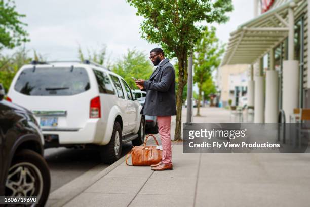 young man looks at phone while standing on a sidewalk downtown - duffel tas stockfoto's en -beelden