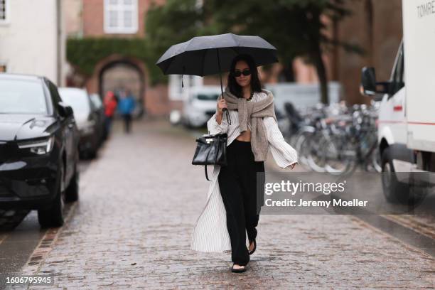 Nisi seen outside Skall Studio show wearing black small sunnies, beige and white striped long linen shirt, Skall Studio knitwear wrapped around...