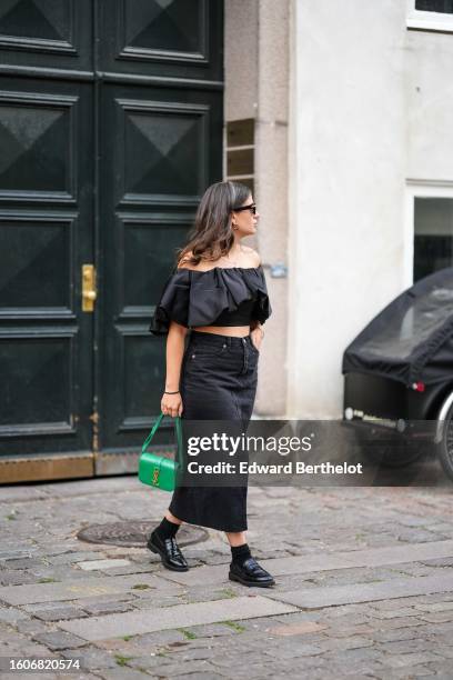 Guest wears black sunglasses, a gold small chain pendant necklace, a black shoulder-off / ruffled short sleeves cropped top, a black denim slit /...